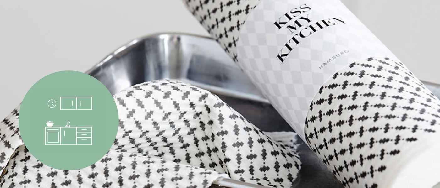 Eine Rolle gemusterter Papierhandtücher mit der Aufschrift „Kiss My Kitchen“ ist teilweise auf einem Metalltablett ausgerollt. Ein grünes rundes Symbol an der Seite zeigt eine vereinfachte Küchenillustration.