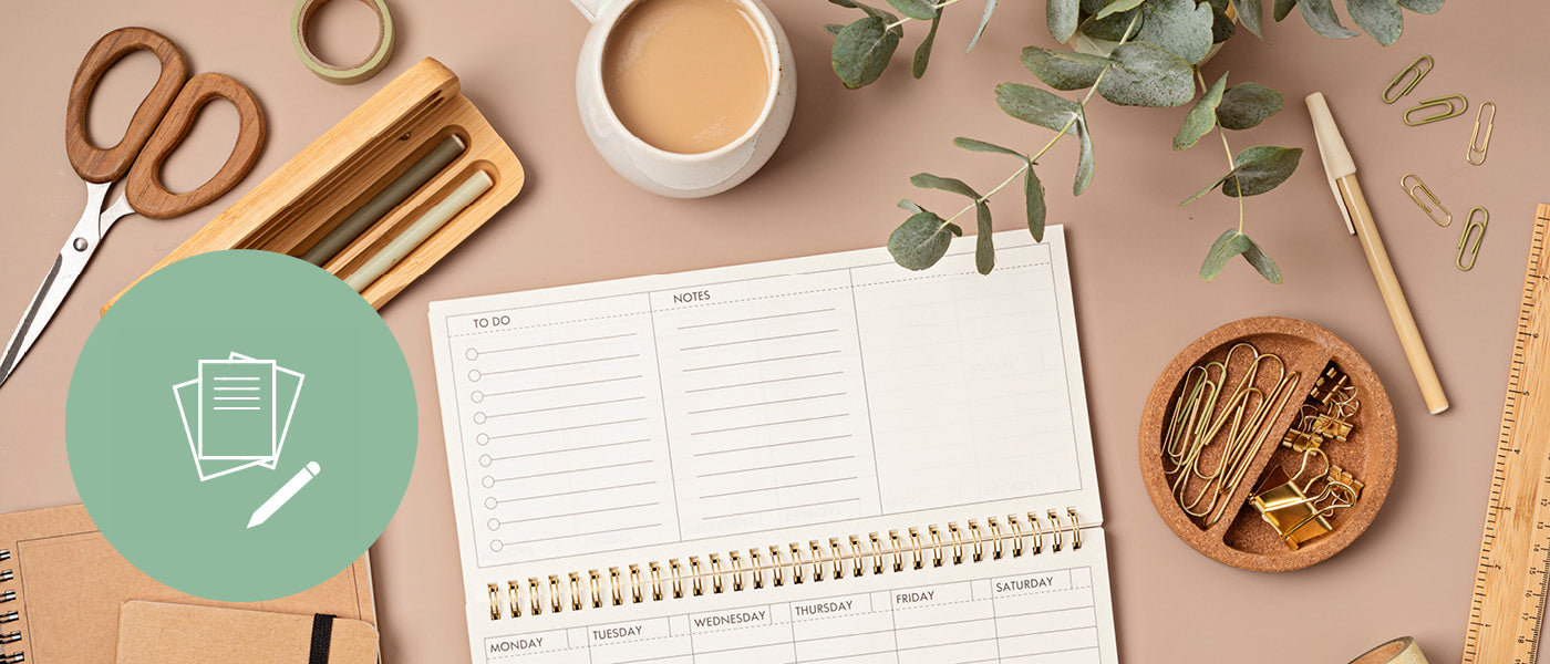 Draufsicht auf einen Planer, der für ein Wochenlayout auf einem Schreibtisch mit einer Kaffeetasse, Schere, Büroklammern, einem Holzlineal und einer Pflanze geöffnet ist. In der Ecke befindet sich ein grüner Kreis mit einem Papier- und Stiftsymbol.