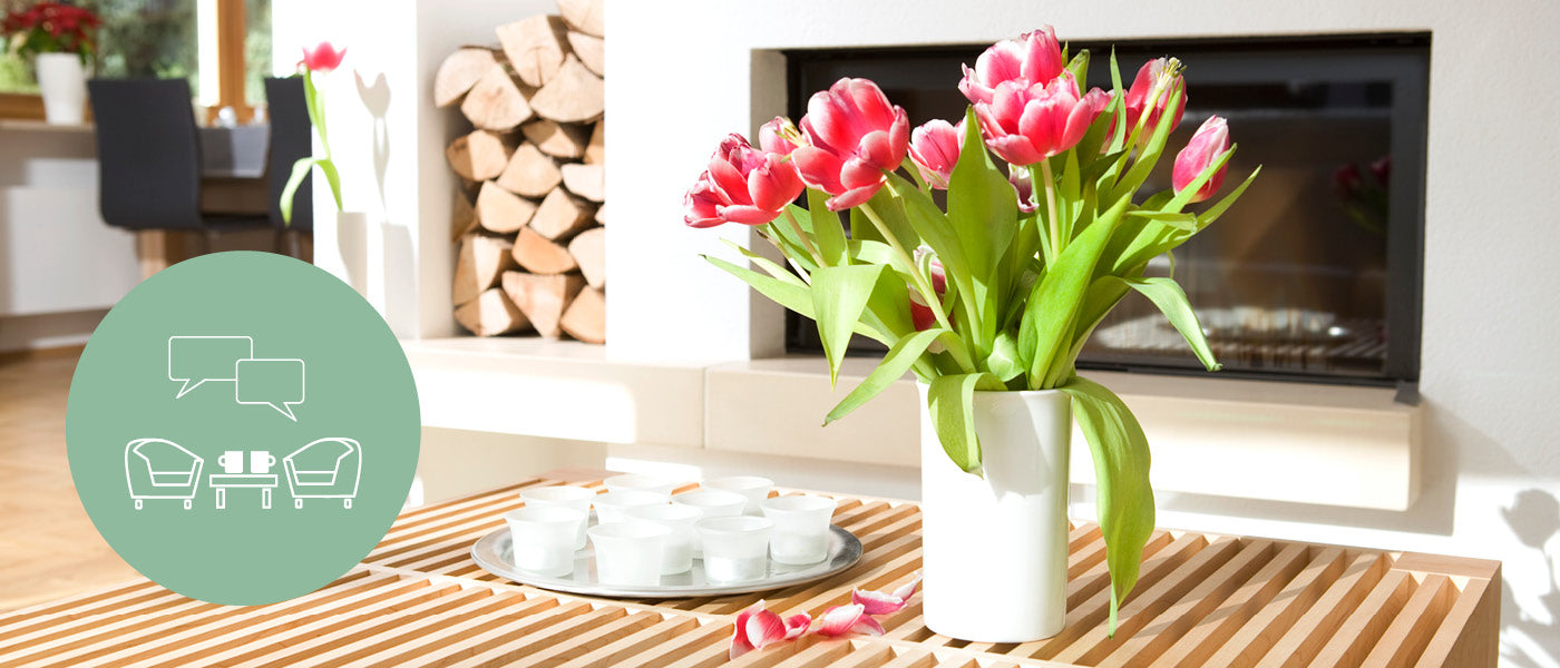 Vase mit roten Tulpen auf einem Holzlattentisch in einem hellen Wohnzimmer mit gestapeltem Brennholz und einem Kamin. Links ein Symbol mit Stühlen, einem Tisch und Sprechblasen auf einem grünen Kreis.