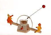 Die Kraul Lichtwippe mit Püppchen von Kraul ist ein Wippspielzeug aus Holz mit zwei orangefarbenen Stofffiguren, einer leuchtenden Teelichtkerze in der Mitte und einem Bimetallstab aus Metall mit einer roten Kugel an der Spitze.