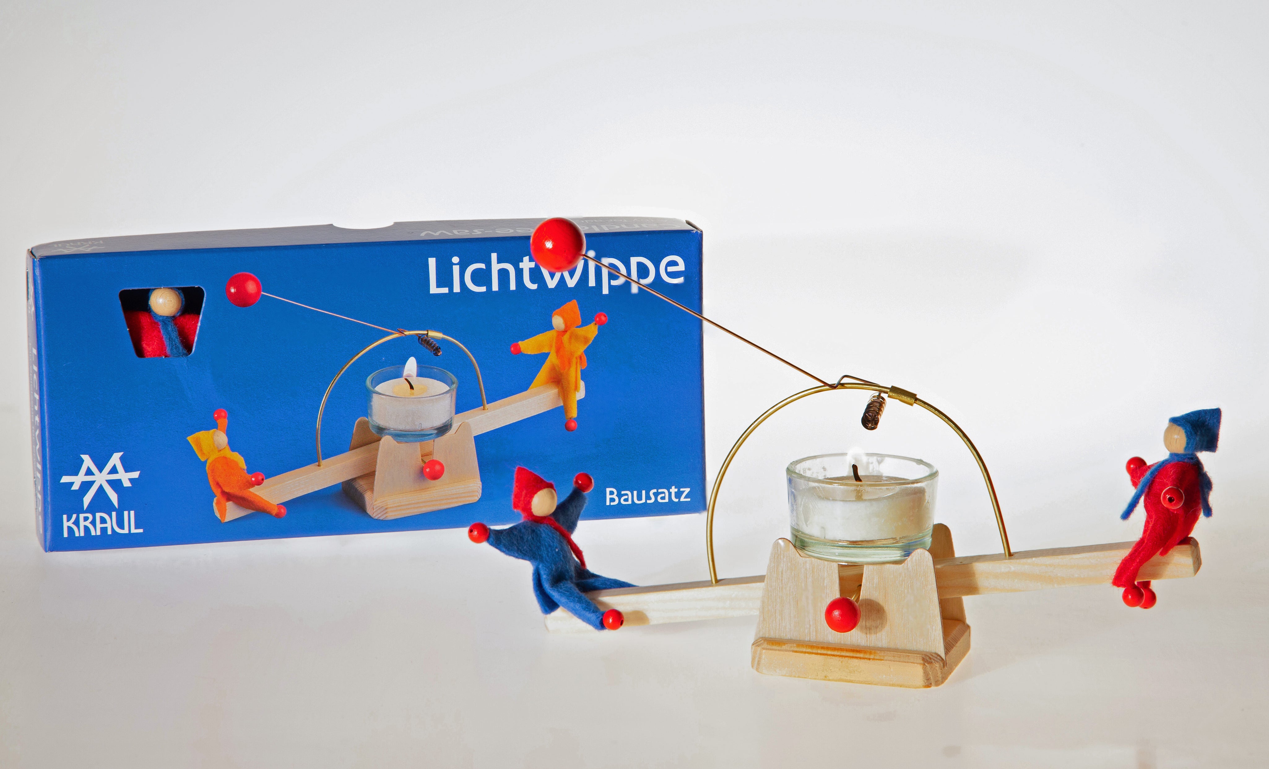 Die Kraul - Lichtwippe mit Püppchen ist eine Holzwippe mit bunten Figuren, die einen kleinen Teelichthalter balanciert. Der Markenname Kraul und ein Bild der zusammengebauten Wippe sind auf der Verpackung der Lichtwippe abgebildet.