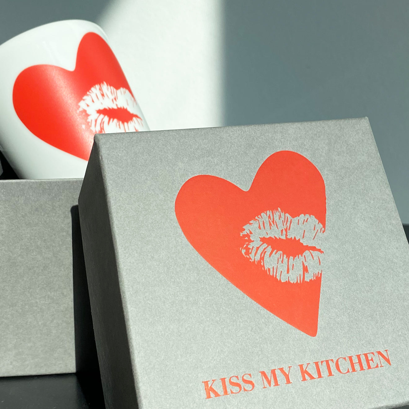 Ein grauer Geschenkkarton mit rotem Herz und Lippenaufdruck und der Aufschrift "KISS MY KITCHEN" steht neben einem buchblauen Porzellanbecher mit rotem Kiss-Aufdruck, der bei hellem Tageslicht teilweise sichtbar ist.