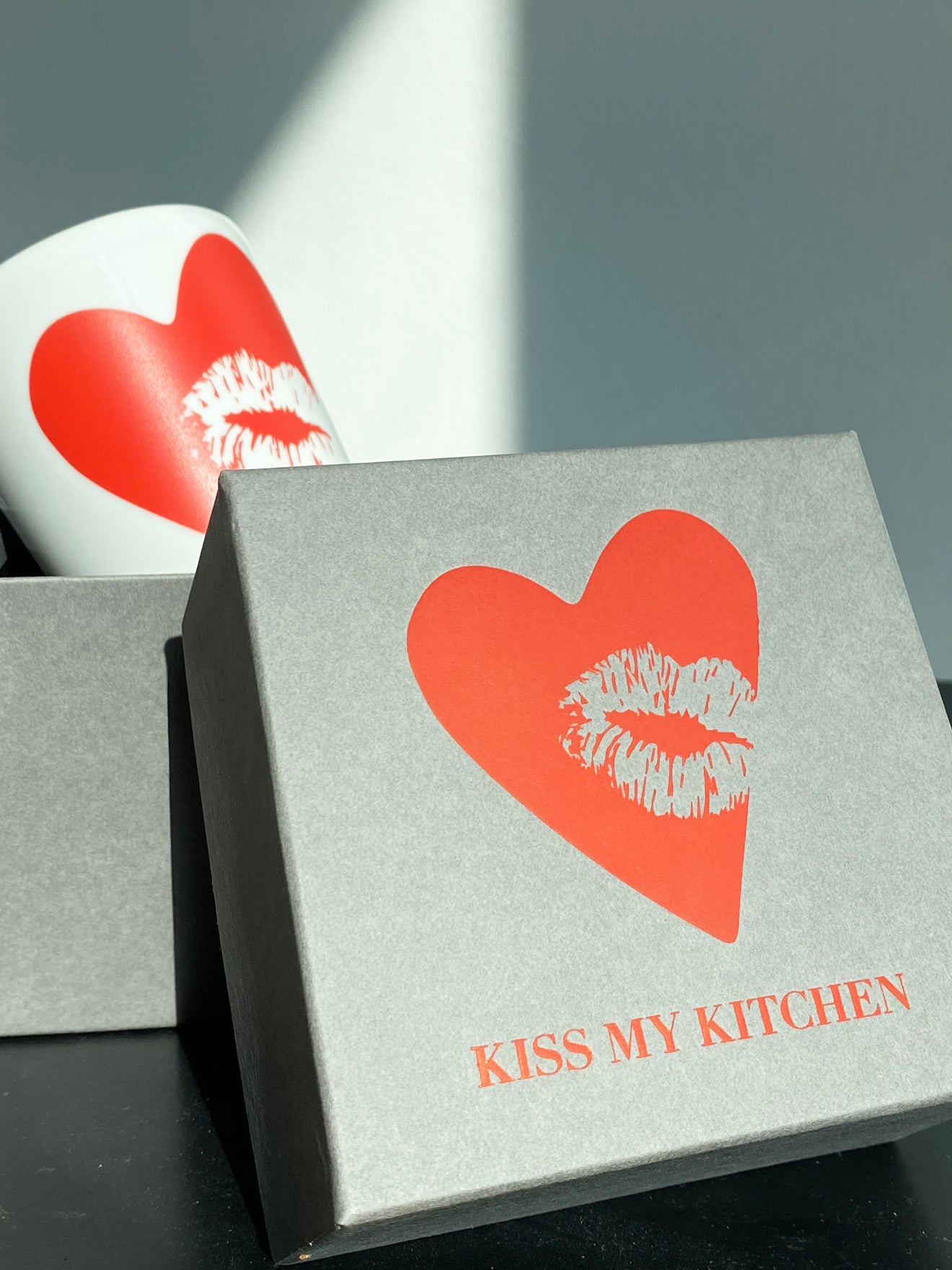Ein grauer Geschenkkarton mit rotem Herz und Lippenaufdruck und der Aufschrift "KISS MY KITCHEN" steht neben einem buchblauen Porzellanbecher mit rotem Kiss-Aufdruck, der bei hellem Tageslicht teilweise sichtbar ist.