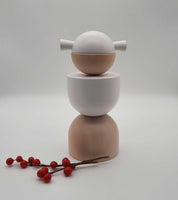 Eine Weihnachtsengel-Skulptur von Cultform präsentiert sich als minimalistischer Stapel glatter, runder Formen in neutralen Farbtönen, gekrönt von einem Engelskopf mit zwei hervorstehenden Flossen. Vor einem schlichten Hintergrund begleiten rote Beeren auf einem Zweig dieses von der Figurenserie inspirierte Erzgebirge-Stück.