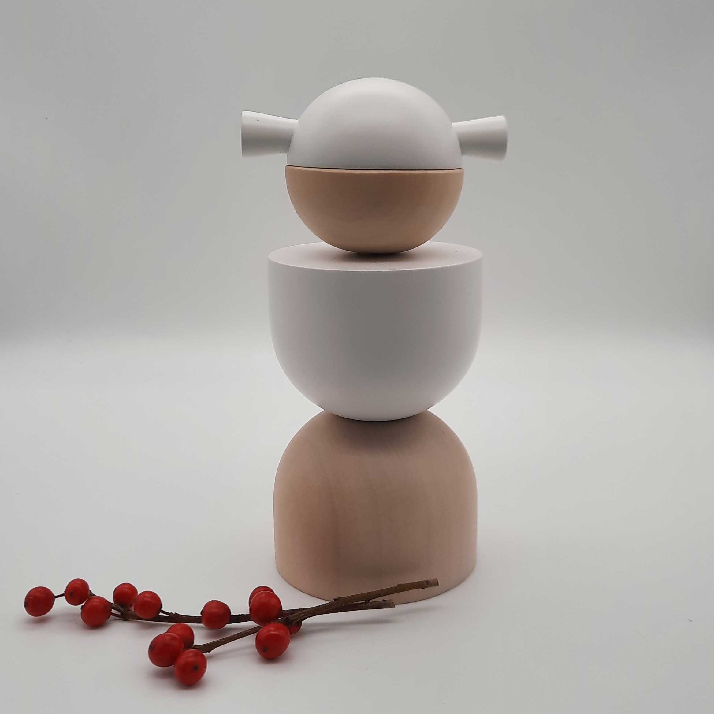 Eine Weihnachtsengel-Skulptur von Cultform präsentiert sich als minimalistischer Stapel glatter, runder Formen in neutralen Farbtönen, gekrönt von einem Engelskopf mit zwei hervorstehenden Flossen. Vor einem schlichten Hintergrund begleiten rote Beeren auf einem Zweig dieses von der Figurenserie inspirierte Erzgebirge-Stück.