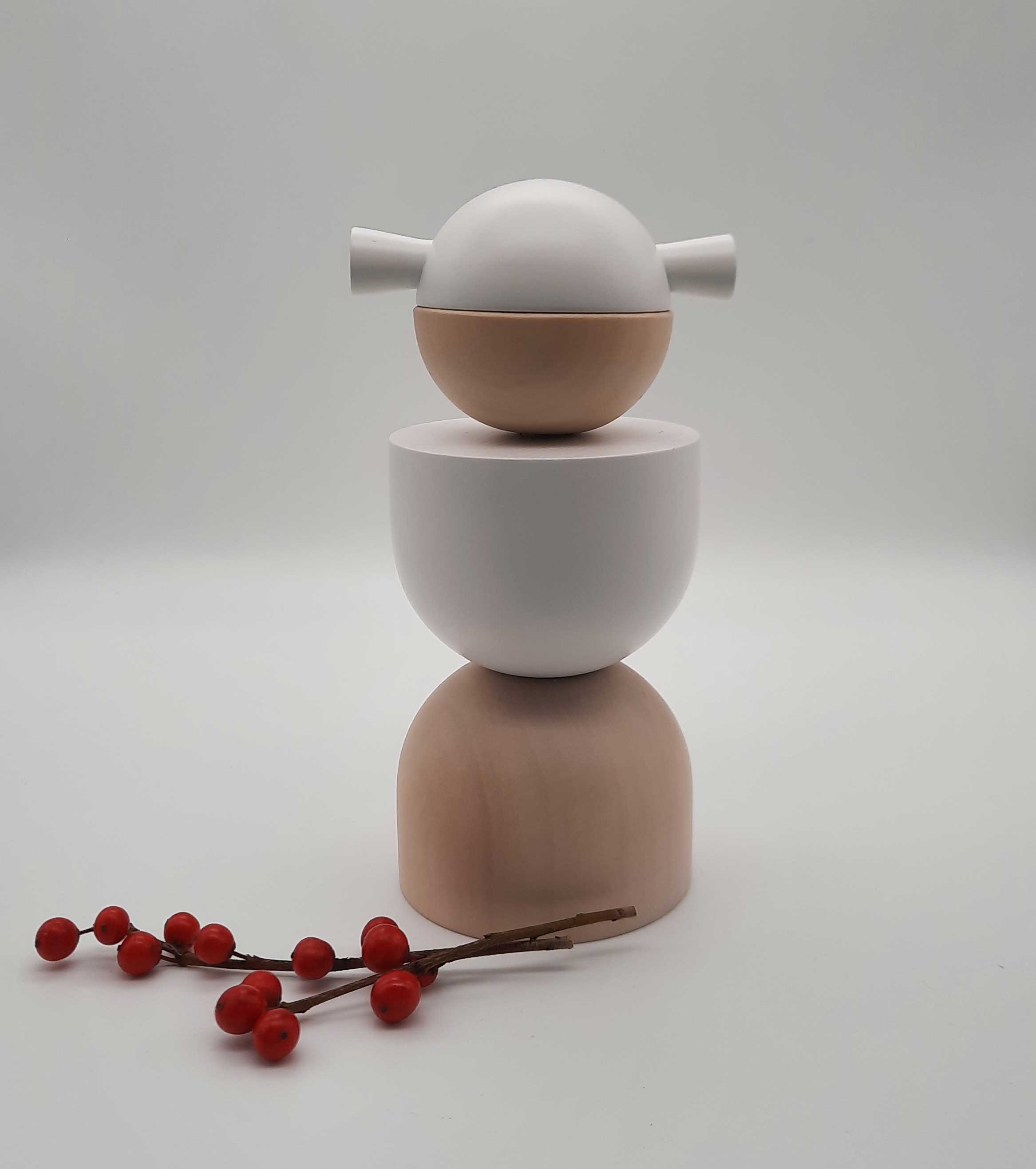 Eine Weihnachtsengel-Skulptur von Cultform präsentiert sich als minimalistischer Stapel glatter, runder Formen in neutralen Farbtönen, gekrönt von einem Engelskopf mit zwei hervorstehenden Flossen. Vor einem schlichten Hintergrund begleiten rote Beeren auf einem Zweig dieses von der Figurenserie inspirierte Erzgebirge-Stück.