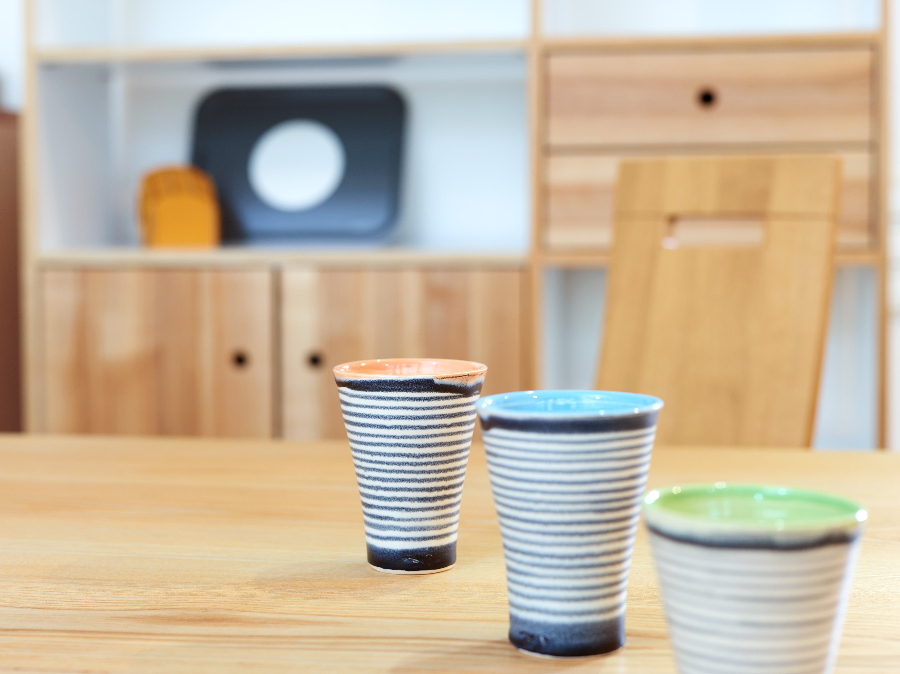 Drei Keramikbecher gestreift von Susanne Protzmann, dünne Keramik mit bunten Rändern (orange, blau und grün), stehen auf einem Holztisch. Im Hintergrund ist ein Holzregal zu sehen, das mit verschiedenen Objekten geschmückt ist, darunter ein schwarzes Tablett und ein kleiner orangefarbener Gegenstand.