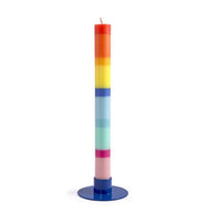Eine hohe, zylindrische „Grußkerze Schulkind“ (nicht das Mädchen), aus Stearinwachs mit horizontalen Regenbogenschichten, steht auf einem runden blauen Sockel vor einem weißen Hintergrund.