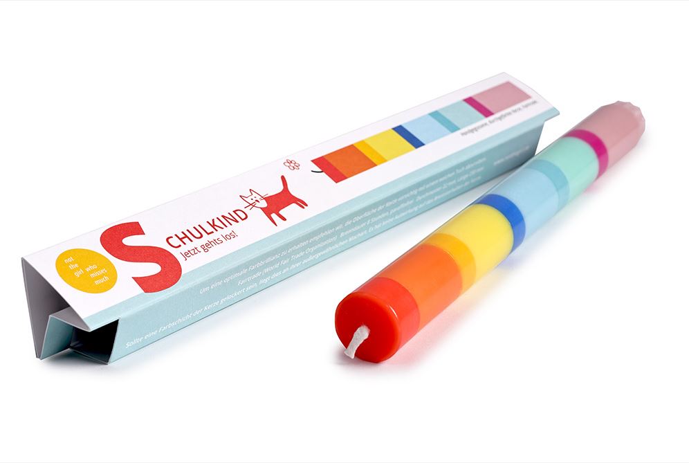 Eine bunte, gestreifte „Grußkerze Schulkind“-Kerze von „not the girl“ mit roten, gelben, blauen und grünen Streifen liegt neben ihrer dreieckigen Schachtel, auf der eine rote Katze und der Markenname auf weißem Hintergrund abgebildet sind.