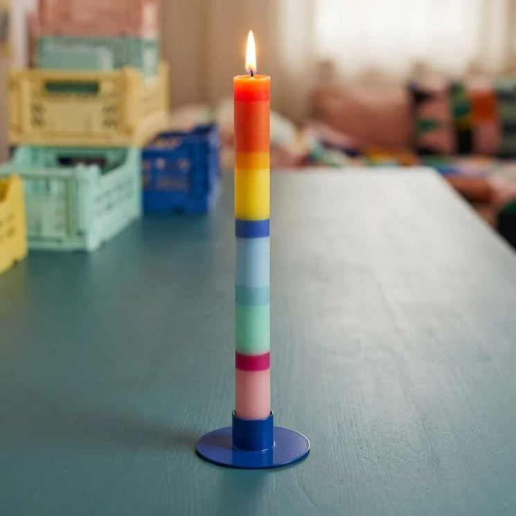 Die „Grußkerze Schulkind“ von „not the girl“, eine hohe vegane Kerze mit horizontalen Regenbogenstreifen, steht in einem blauen Halter auf einem blaugrünen Tisch, im Hintergrund sind bunte Kisten und ein verschwommener, gemütlicher Raum zu sehen.