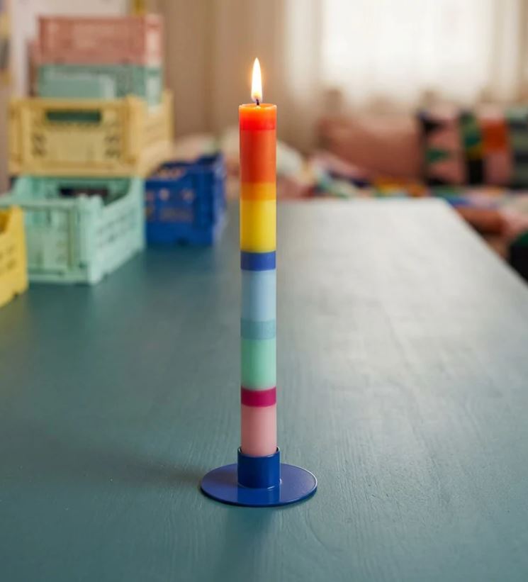 Die „Grußkerze Schulkind“ von „not the girl“, eine hohe vegane Kerze mit horizontalen Regenbogenstreifen, steht in einem blauen Halter auf einem blaugrünen Tisch, im Hintergrund sind bunte Kisten und ein verschwommener, gemütlicher Raum zu sehen.