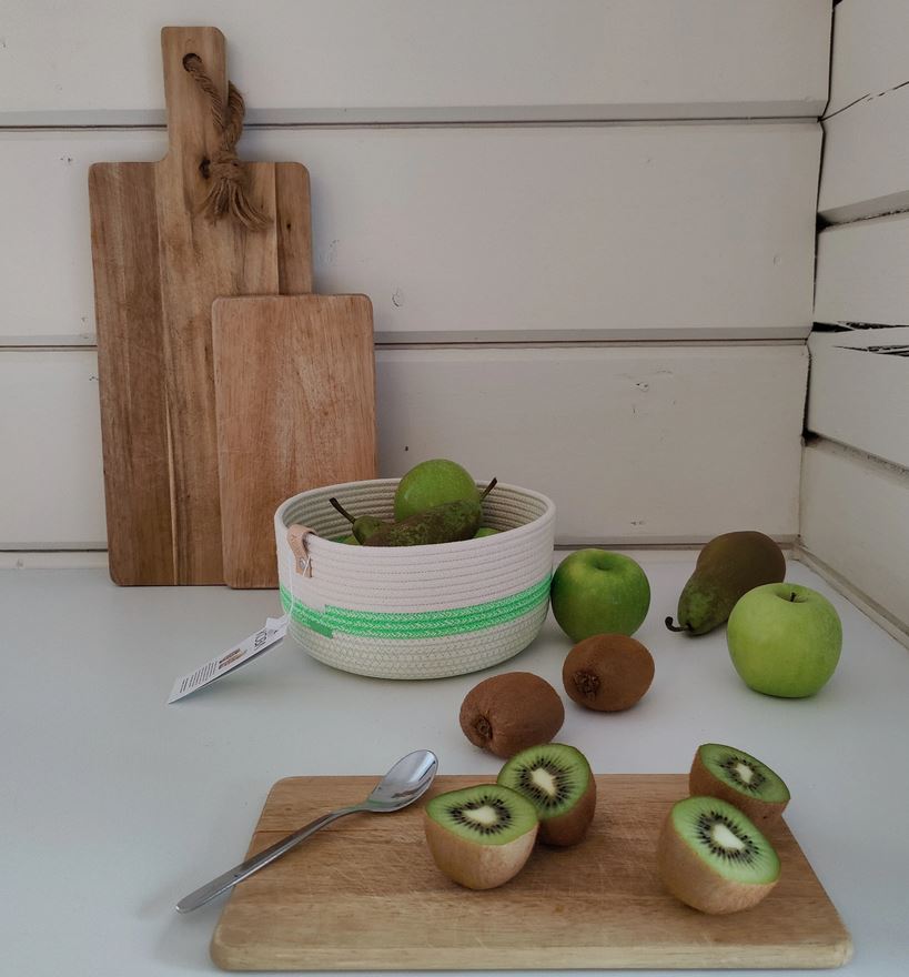 Eine Küchenszene mit einem hellen Holzschneidebrett, auf dem geschnittene und ganze Kiwis liegen. Daneben liegt ein Löffel. Im Hintergrund liegen grüne Äpfel und Birnen in einer Koba Korb Koba Bowl Fluo Green aus recyceltem Baumwollkordel mit grünem Streifen. Zwei Holzschneidebretter sind an eine Wand gelehnt.