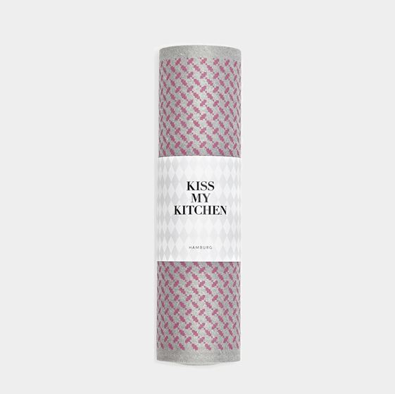 Eine aufgerollte Kiss My Kitchen Schwammtuchrolle in Pali-Rosa-Grau mit rosa Rautenmuster ist in ein weißes Etikett mit der Aufschrift „KISS MY KITCHEN HAMBURG“ eingewickelt. Das kompostierbare Tuch liegt auf einem schlichten hellgrauen Untergrund.