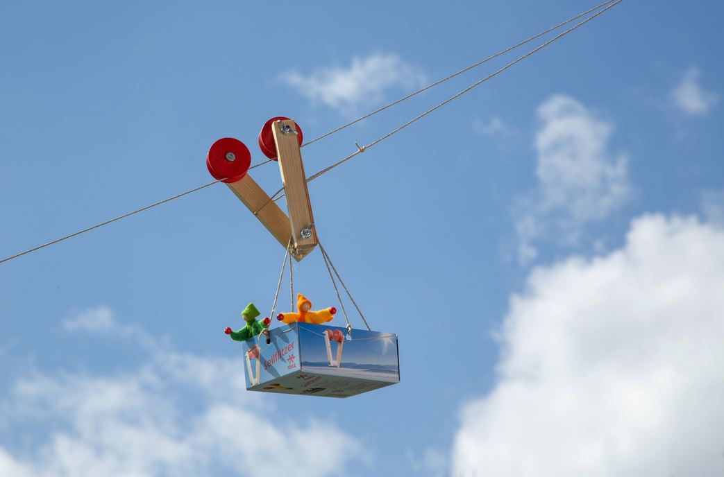 Zwei Plüschtiere fahren in einer kleinen Box auf der Kraul - Seilflitzer Seilrutsche von Kraul und genießen den Nervenkitzel des Seilbahnspaßes unter einem blauen Himmel mit vereinzelten Wolken.