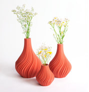 Vor einem weißen Hintergrund sind drei Terrakotta-Vasen „Swirl“ S von nevalu zu sehen, die eine orangefarbene, strukturierte Keramikoptik aufweisen und aus recycelten Materialien gefertigt sind. Jede Vase zeigt zarte weiße und gelbe Blumen, die der Szene eine frische, natürliche Note verleihen und ein nachhaltiges Designethos verkörpern.