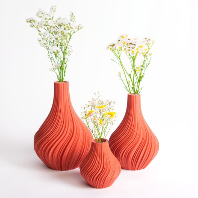 Vor einem weißen Hintergrund sind drei Terrakotta-Vasen „Swirl“ S von nevalu zu sehen, die eine orangefarbene, strukturierte Keramikoptik aufweisen und aus recycelten Materialien gefertigt sind. Jede Vase zeigt zarte weiße und gelbe Blumen, die der Szene eine frische, natürliche Note verleihen und ein nachhaltiges Designethos verkörpern.