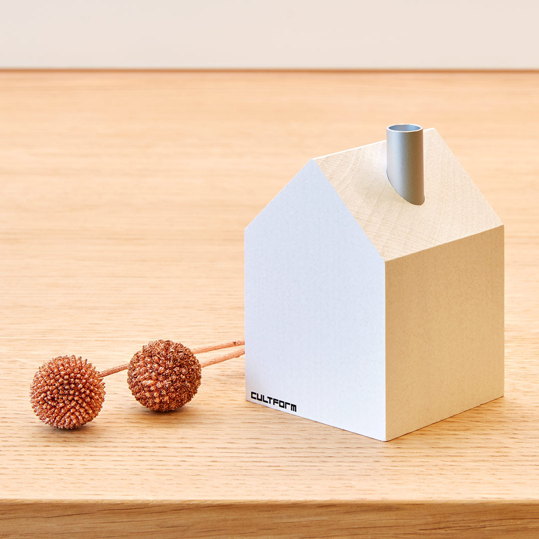 Ein minimalistisches weißes, hausförmiges Objekt mit einem Schornstein steht auf einer Holzoberfläche und erinnert an den Charme traditioneller Räucherhäuser aus dem Erzgebirge. Flankiert wird es von zwei braunen Kugeln mit Textur. Die Marke „cultform“ ist auf diesem exquisiten Stück elegant abgebildet und deutet darauf hin, dass es für einen Designpreis geeignet ist.