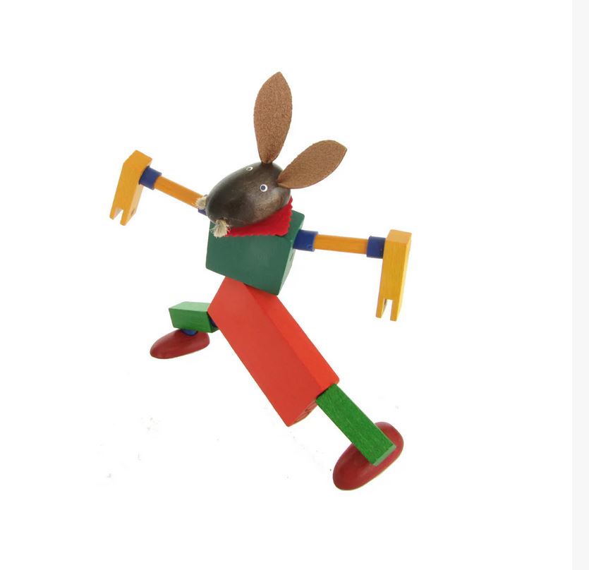 Ein buntes buchenblaues Häslein Hopps - Kellner Steckfigur mit Hasenkopf, roter Hose, grünem Hemd, großen braunen Ohren, ausgestreckten Armen und roten Schuhen nimmt eine verspielte Pose auf weißem Hintergrund ein.