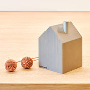 Ein kleines, minimalistisches Objekt in Form eines Holzhauses aus hochwertigem Holz mit einem zylindrischen Vorsprung an der Oberseite. Es ruht auf einer Holzoberfläche neben zwei strukturierten, kugelförmigen braunen Objekten, die links durch eine Schnur verbunden sind. Dieses Stück erinnert an Räucherhäuser aus dem Erzgebirge und trägt an seiner Seite das Wort „cultform“. Das Produkt ist als Minimalistisches Räucherhaus in Grau von cultform bekannt.