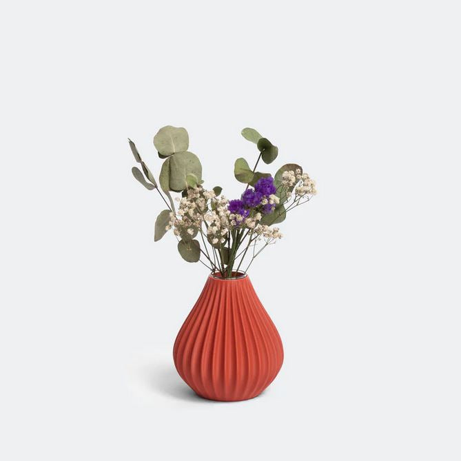 Eine gerippte Terrakotta-Regentropfenvase von nevalu, gefüllt mit einem gemischten Blumenarrangement. Der Strauß umfasst Eukalyptusblätter, Schleierkraut und kleine violette Blüten. Die Vase besteht aus recyceltem Kunststoff und steht auf einem schlichten, hellen Hintergrund, was ihre lebendige Farbe und ihr Engagement für nachhaltiges Design betont.