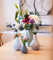 Vier Nevalu-Regentropfenvasen aus Terrakotta, jede mit verschiedenen Blumen wie Lilien, Chrysanthemen und Disteln, stehen auf einem Holztisch. Der Hintergrund zeigt ein verschwommenes Interieur mit minimalistischem Dekor.