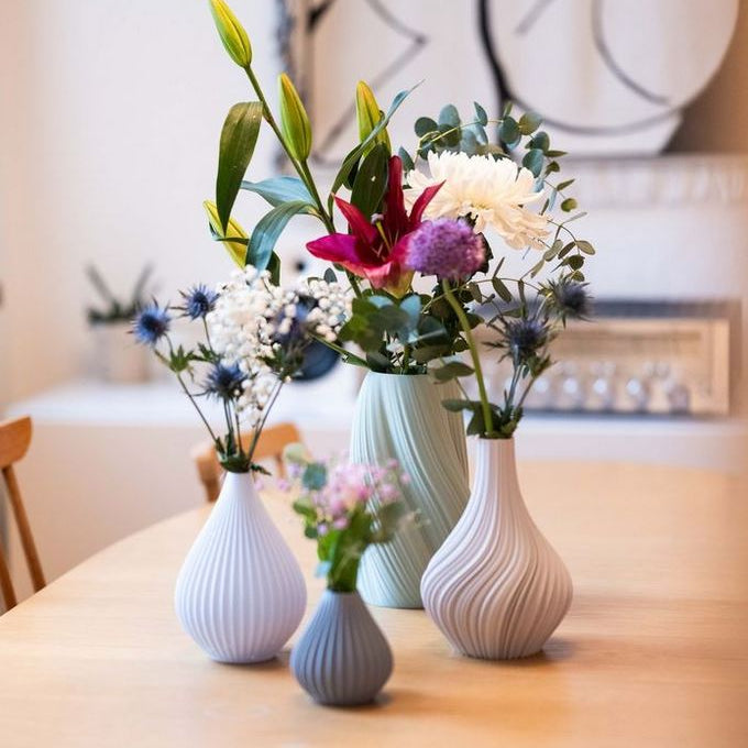 Vier Nevalu-Regentropfenvasen aus Terrakotta, jede mit verschiedenen Blumen wie Lilien, Chrysanthemen und Disteln, stehen auf einem Holztisch. Der Hintergrund zeigt ein verschwommenes Interieur mit minimalistischem Dekor.